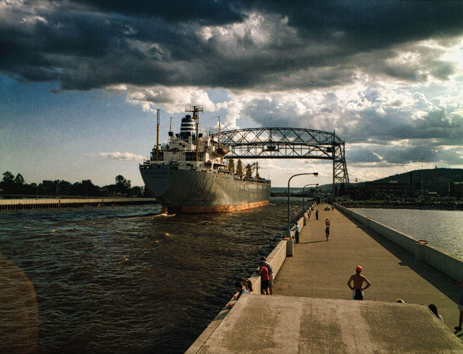 A lake freighter passing under the Aerial Lift Bridge in the Duluth ship canal as people walk the pier, one of the classic things to do in Minnesota